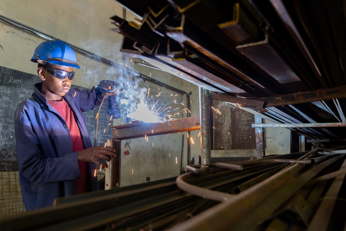A trainee in protective gear welding a steel frame, sparks flying